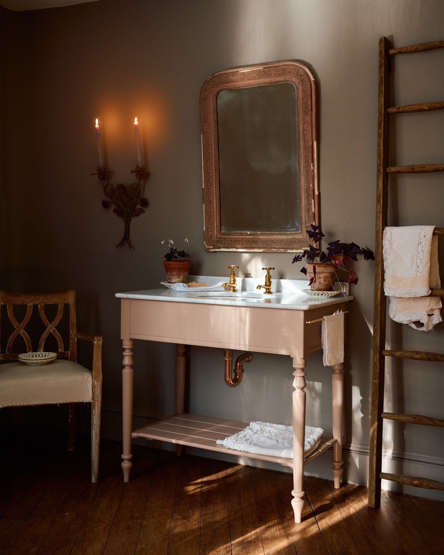 An oblique view towards a dusty-pink Victorian-inspired washstand with brass pillar taps in a greyish-brown painted room. The shot is atmospheric and moody, with two lit candles casting a subtle glow and light from an adjacent window illuminating the brass accents.