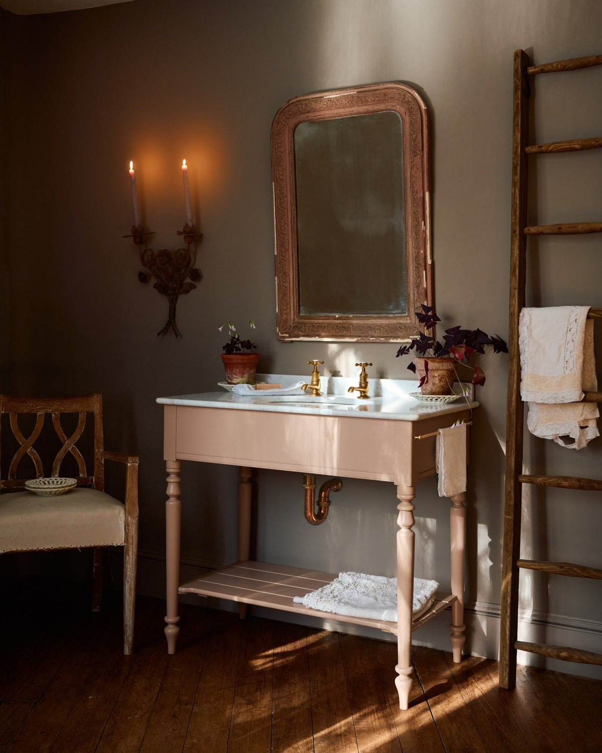 An oblique view towards a dusty-pink Victorian-inspired washstand with brass pillar taps in a greyish-brown painted room. The shot is atmospheric and moody, with two lit candles casting a subtle glow and light from an adjacent window illuminating the brass accents. TEST