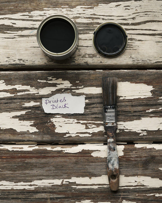An overhead view of an open paint pot and a worn brush on a rustic tabletop. The black colour inside is labelled 'Printer's Black'.