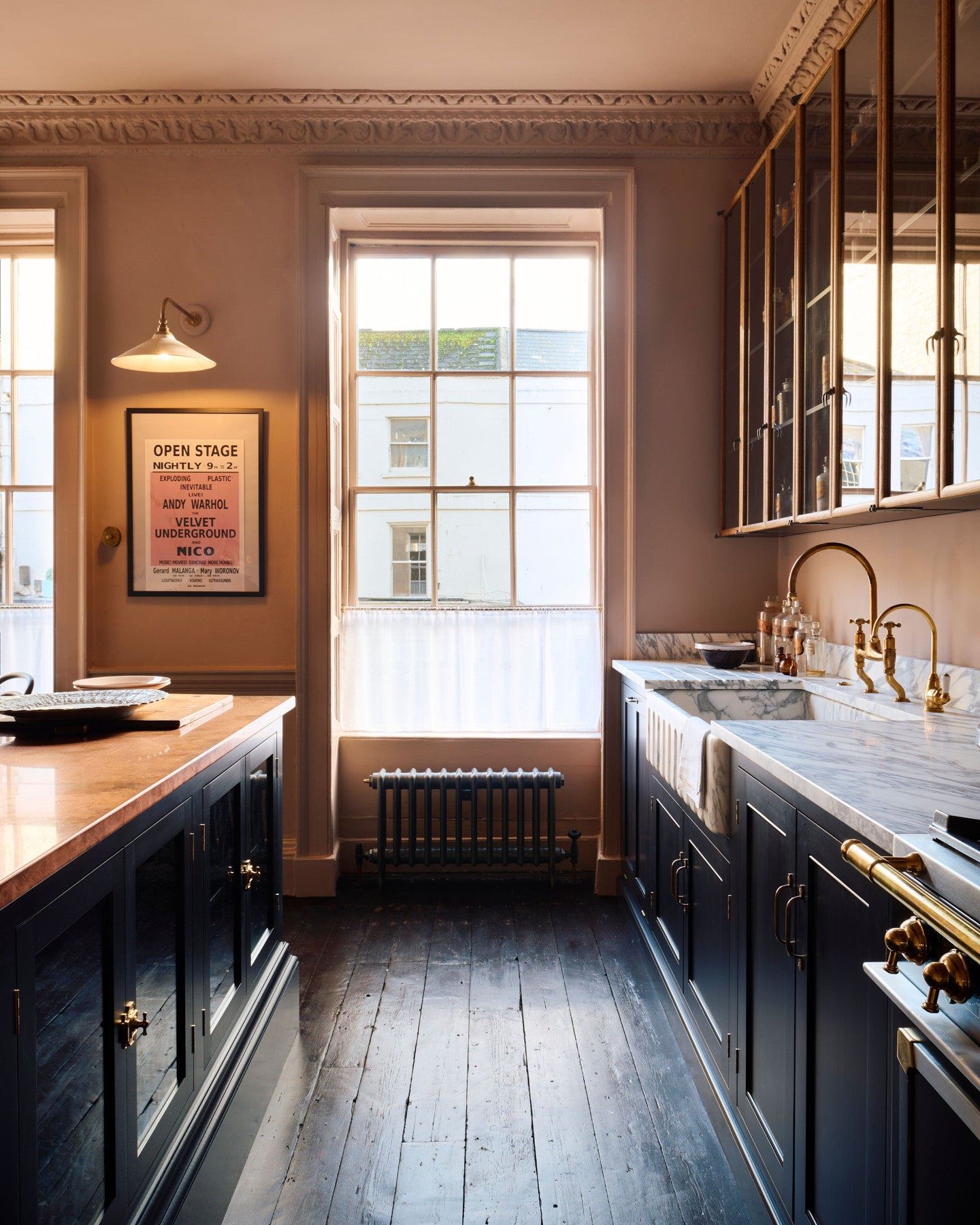 Looking between an island and Georgian-style kitchen sink cabinet with black painted cabinetry, an Arabescato marble sink, an aged brass tap, dark floorboards, pink walls and large original sash window.