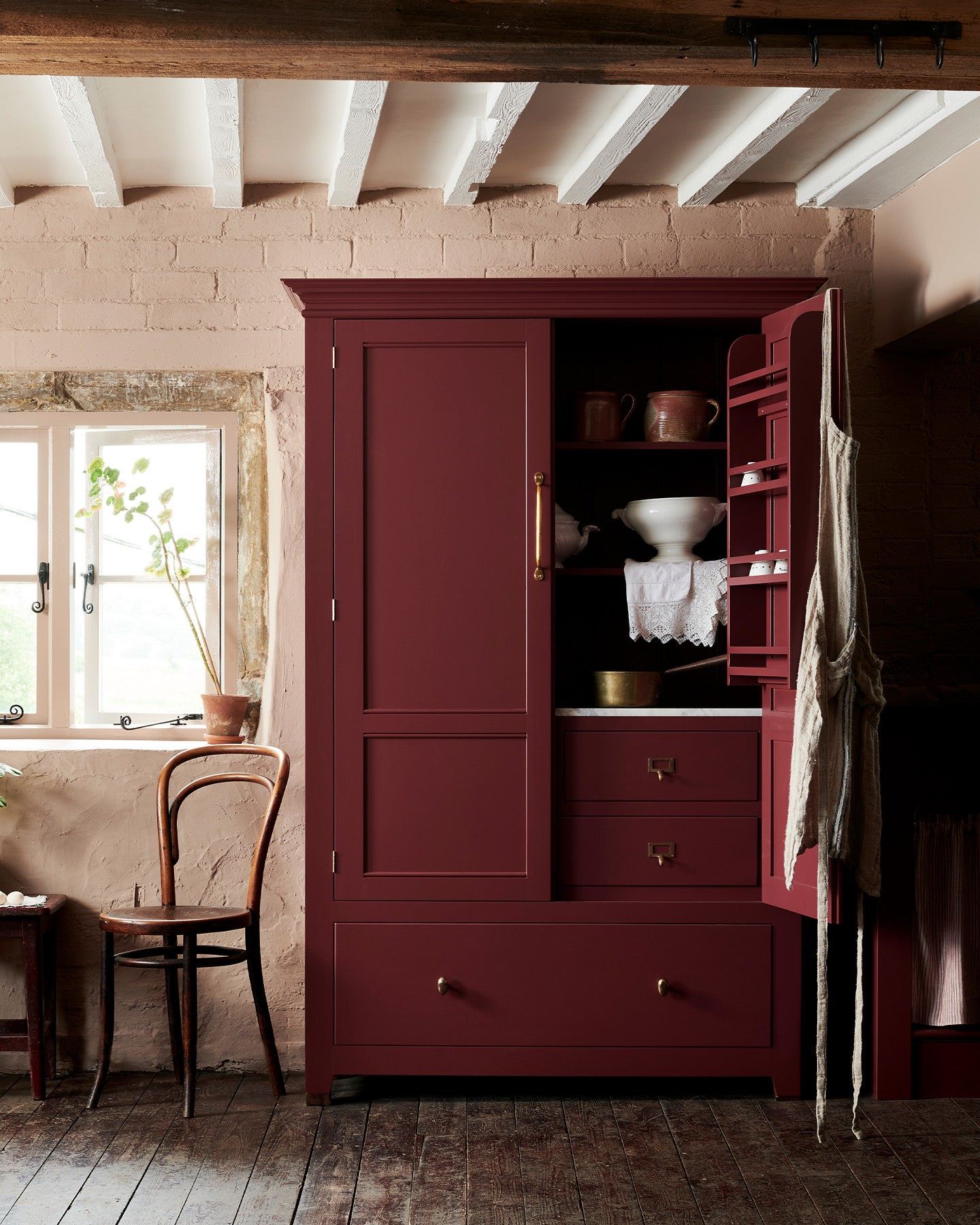 A red painted Georgian-style pantry cupboard with brass hardware and one door open, revealing white vintage crockery inside.