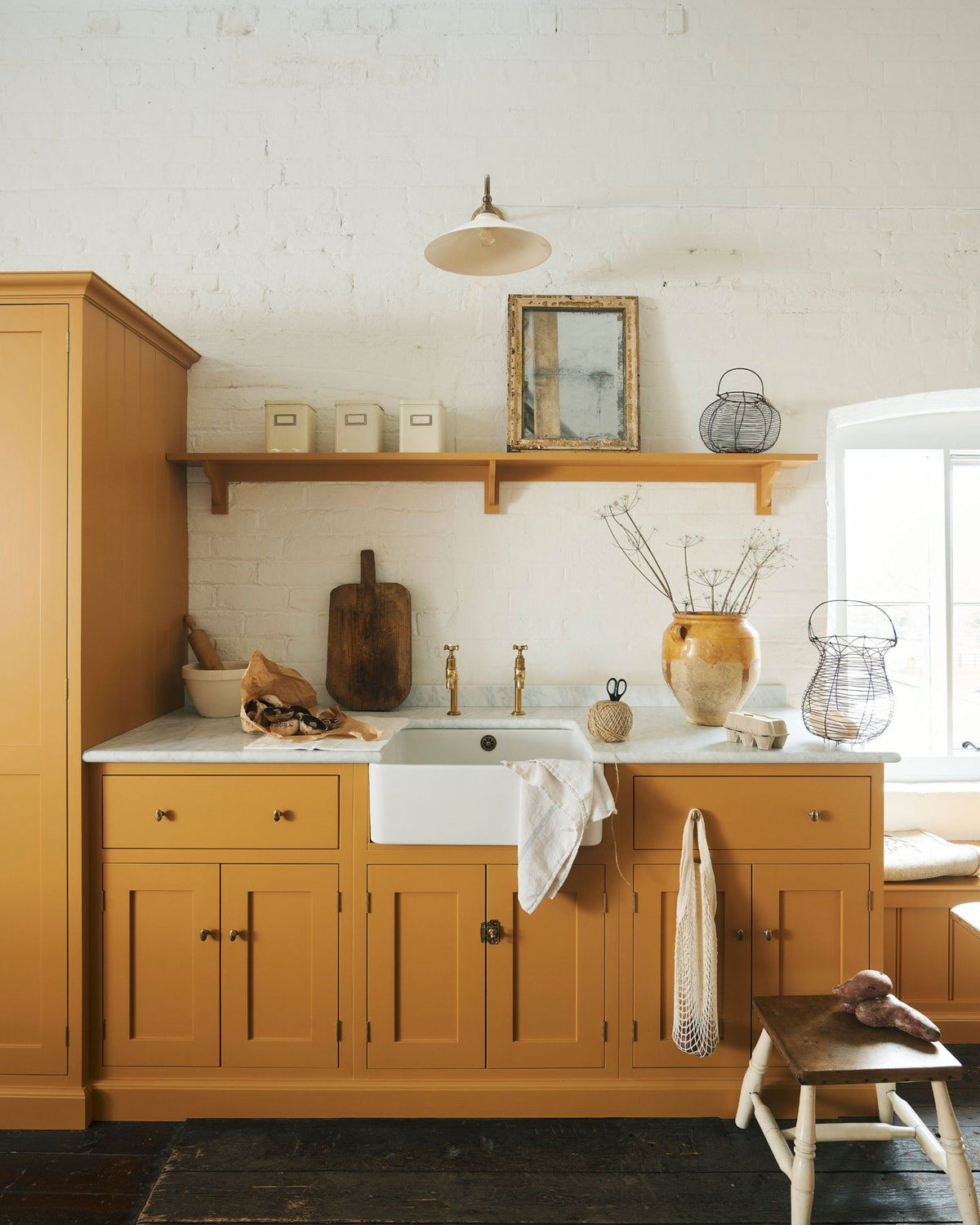A straight-on view of a yellow painted Shaker-style kitchen in a rustic cottage kitchen with dark worn floorboards, white brick walls and wooden beams. TEST