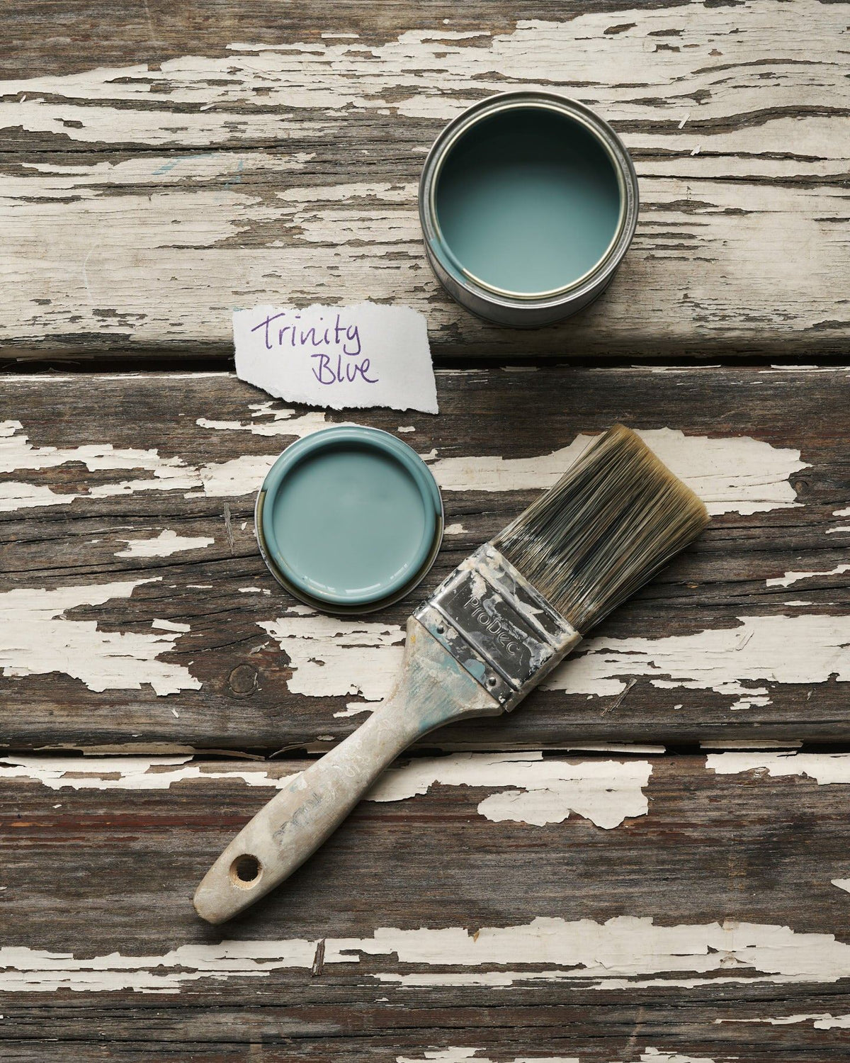 An overhead view of an open paint pot and a worn brush on a rustic tabletop. The duck-egg blue colour inside is labelled 'Trinity Blue'. TEST