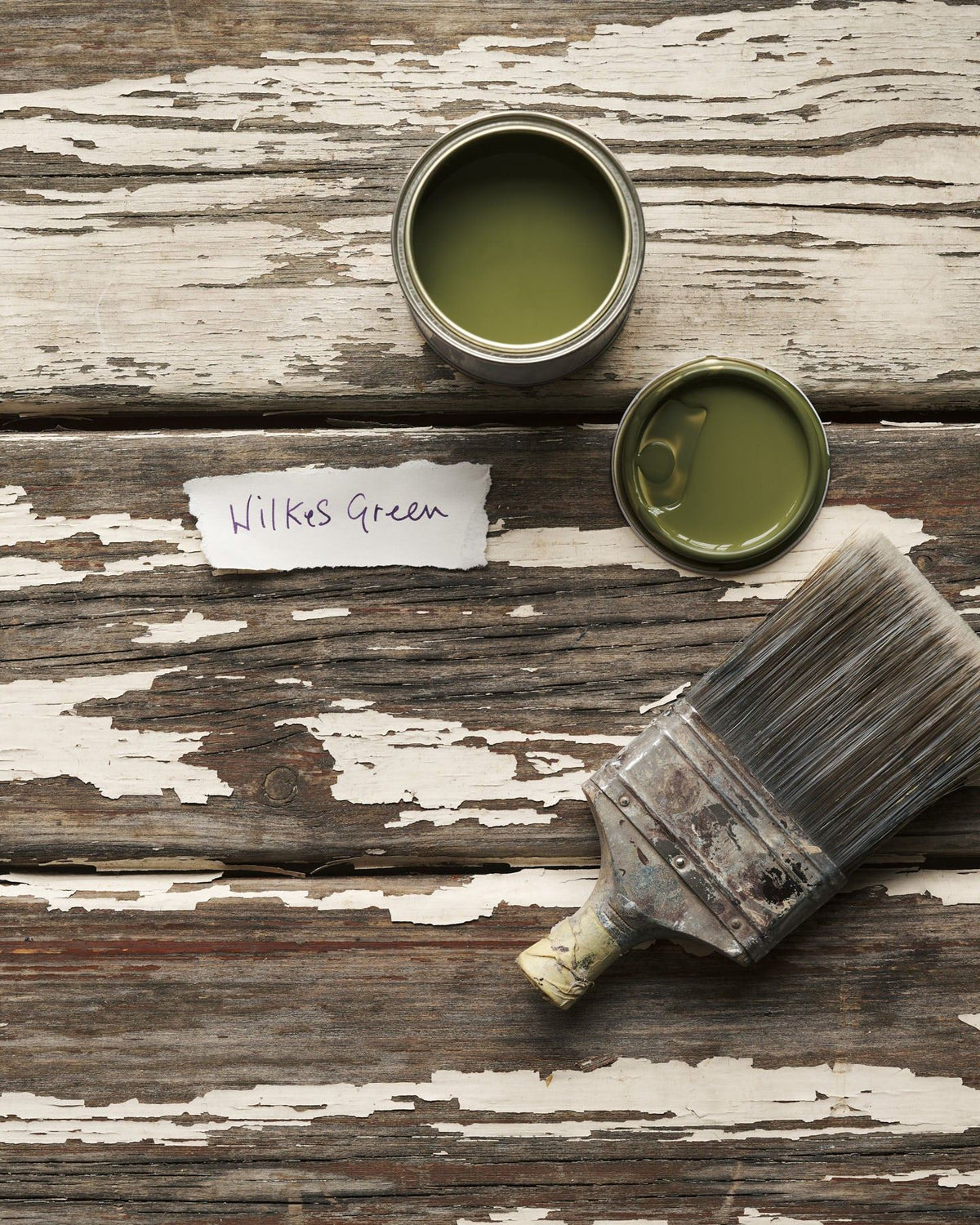 An overhead view of an open paint pot and a worn brush on a rustic tabletop. The mossy green colour inside is labelled 'Wilkes Green'. TEST