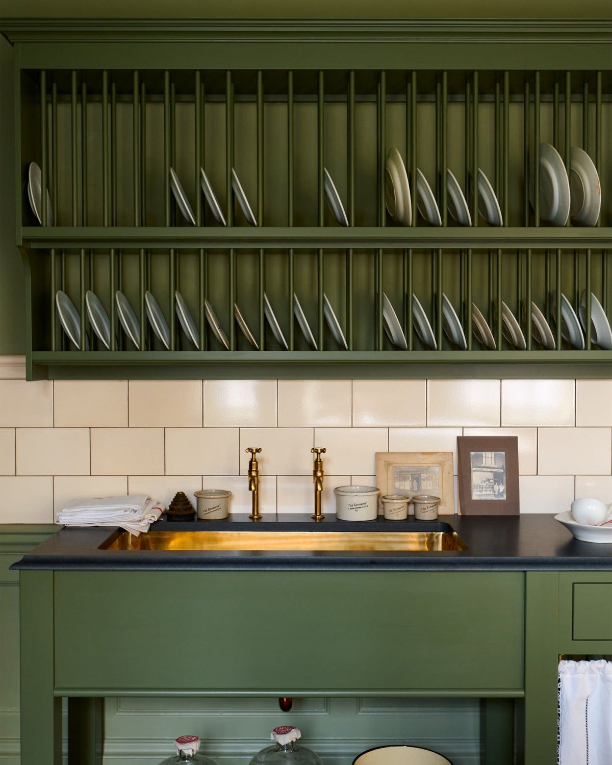 A close-up straight-on view of a mossy green painted Victorian-style sink cupboard with a polished brass sink, aged brass bibcock taps and cream subway wall tiles below a traditional wall-mounted plate rack. TEST
