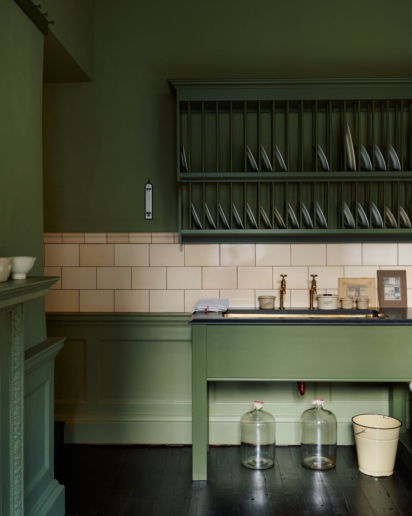 A straight-on view of a mossy green painted Victorian-style sink cupboard with a polished brass sink, aged brass bibcock taps and cream subway wall tiles below a traditional wall-mounted plate rack.