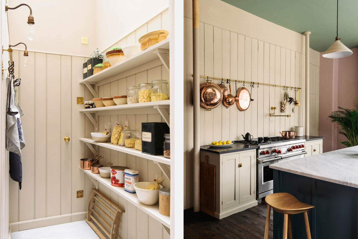 An oblique view of a beige cooker run with a black granite worktop, a silver gas cooker, an aged brass hanging rail, an aged brass pot filler, a pantry room with four long Shaker shelves, and a long kitchen island with a Carrara marble worktop. Lovingly decorated with hanging copper pots and pans, fresh lemons, garlic, chilli and sage, this space balances functionality and style.