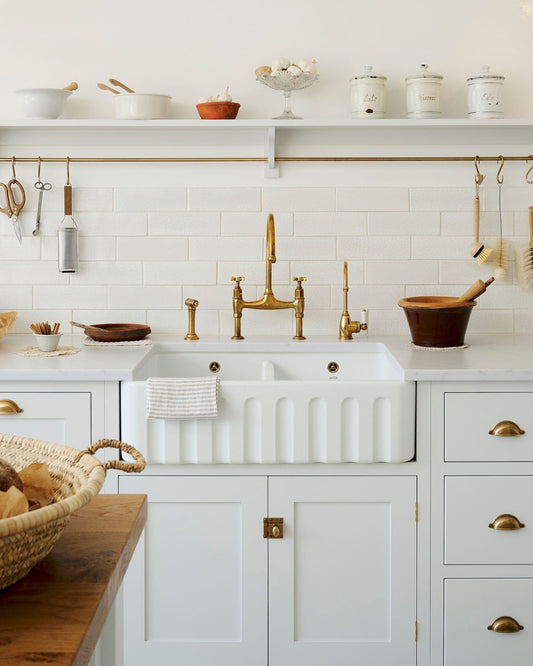 A straight-on view of a large fluted double bowl ceramic Belfast sink in a white painted simple Shaker-style kitchen with an aged brass mixer tap and white metro wall tiles.