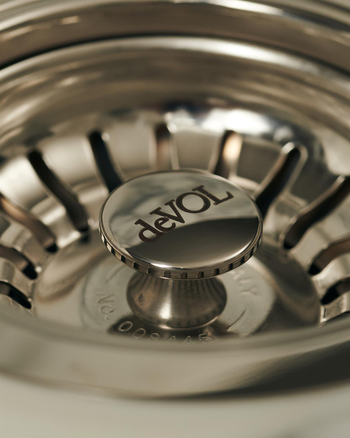 A close-up view of the central finial of a polished chrome basket waste strainer, there is a small 'deVOL' logo in the middle of the finial. TEST