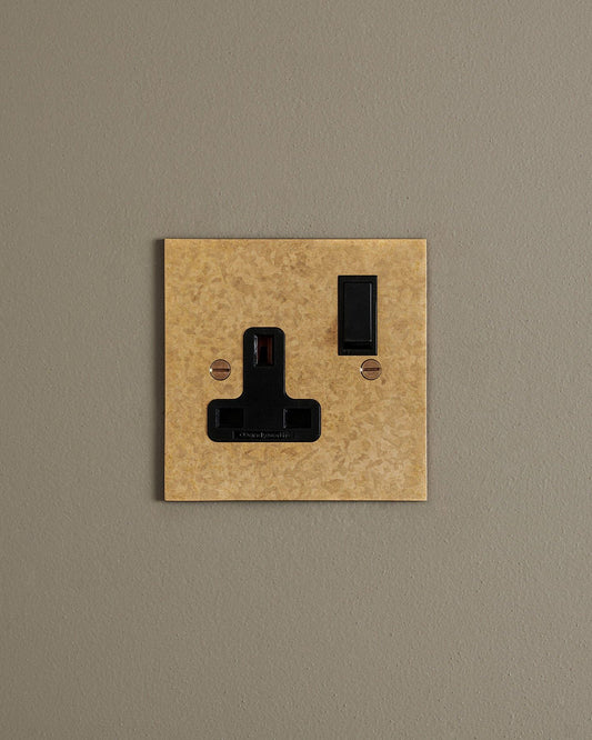 A straight-on view of a classic 13 amp single plug socket in a mottled aged brass finish with black plastic switches.