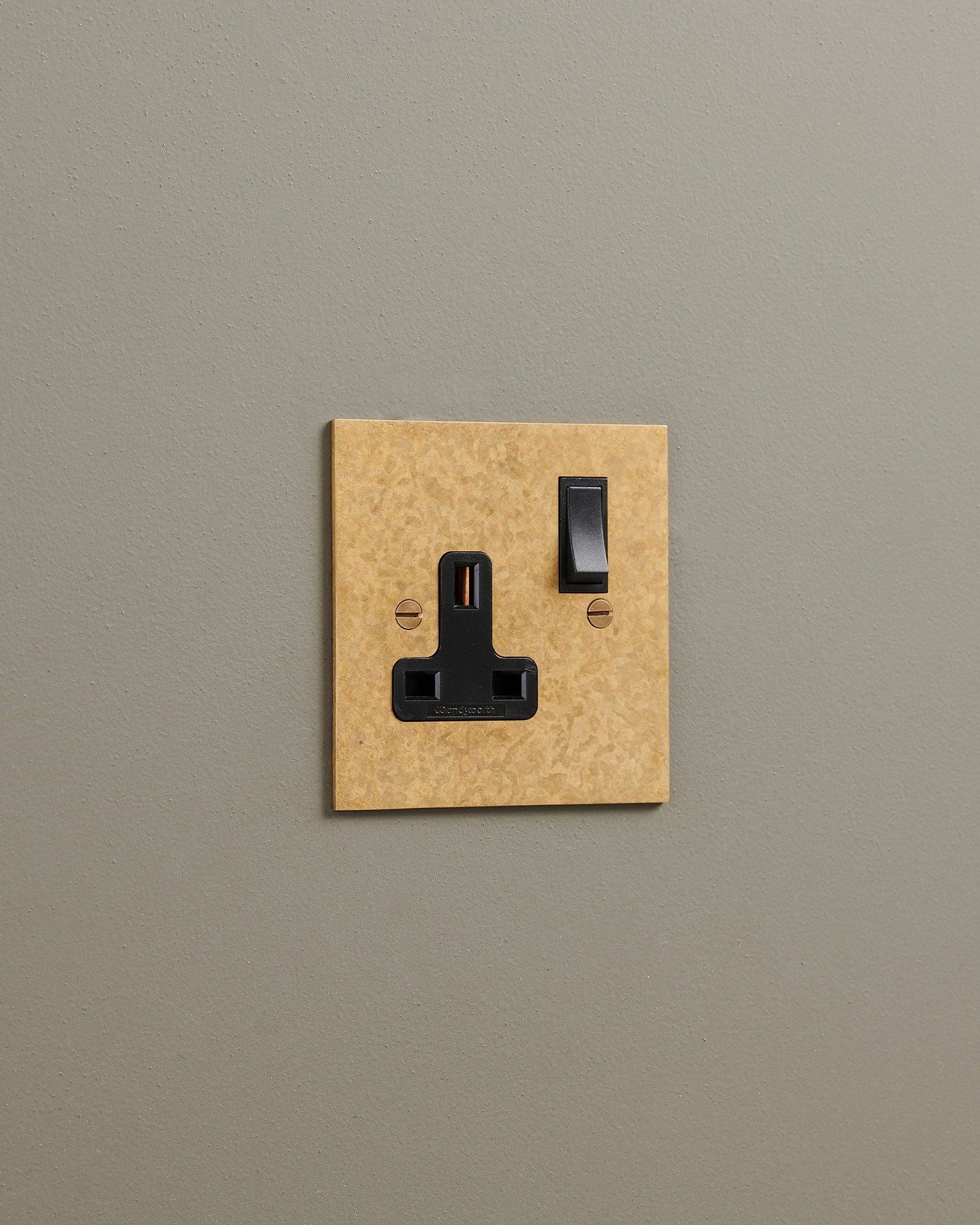 An oblique view of a classic 13 amp single plug socket in a mottled aged brass finish with black plastic switches.
