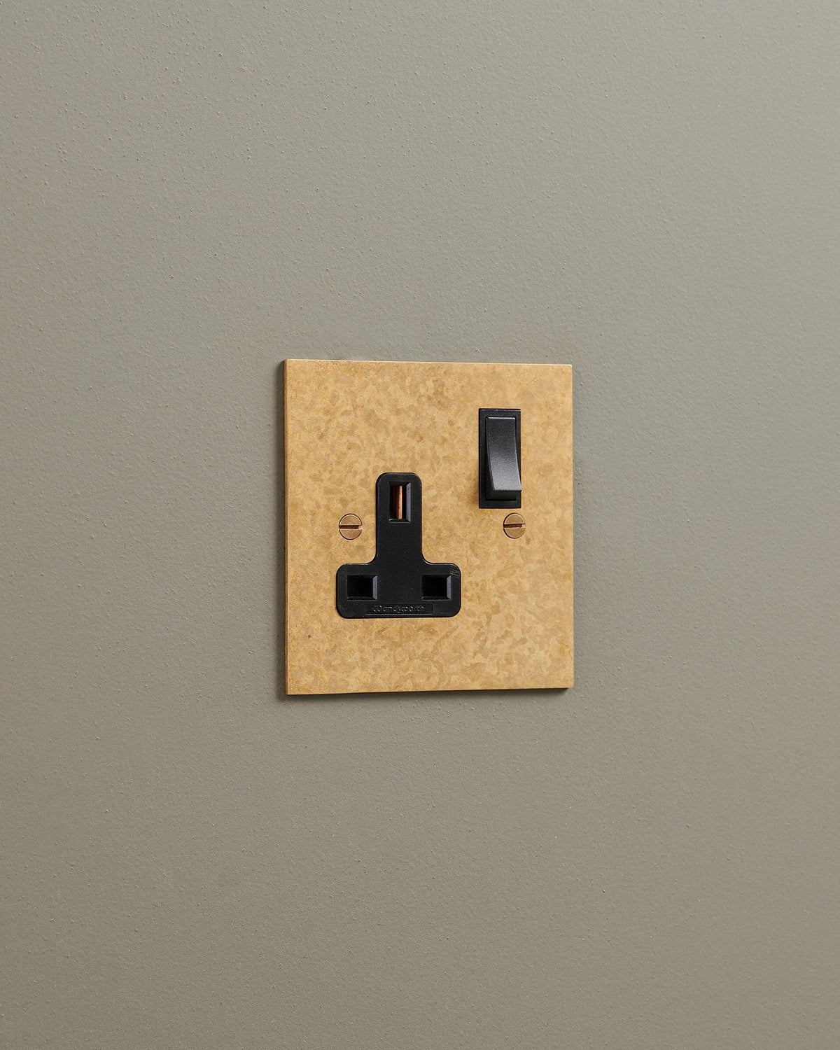 An oblique view of a classic 13 amp single plug socket in a mottled aged brass finish with black plastic switches. TEST