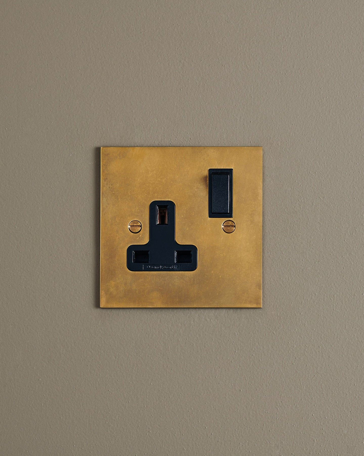 A straight-on view of a classic 13 amp single plug socket in a warm burnished brass finish with black plastic switches. TEST