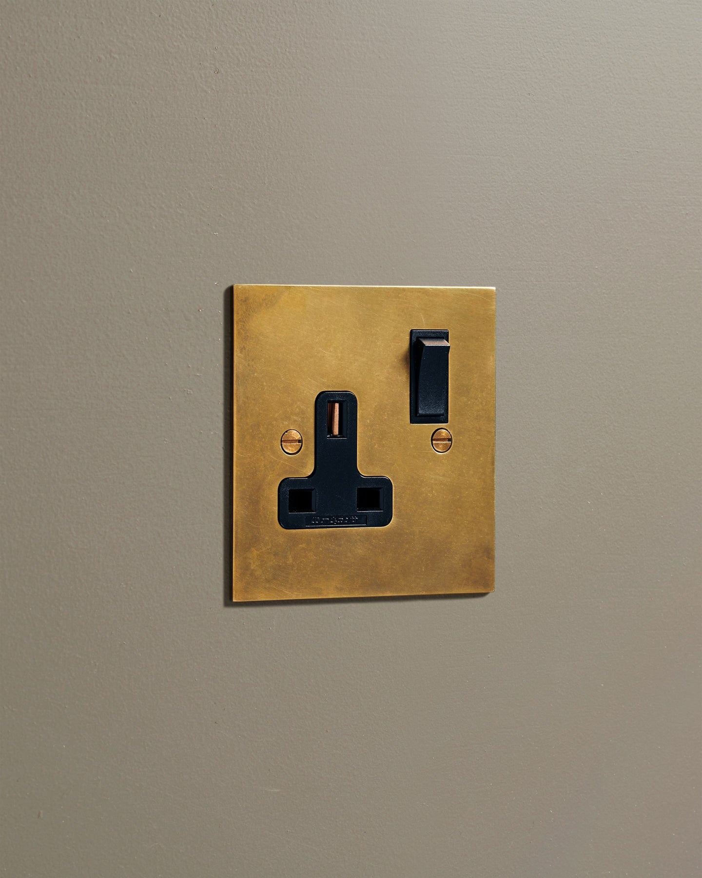 An oblique view of a classic 13 amp single plug socket in a warm burnished brass finish with black plastic switches.