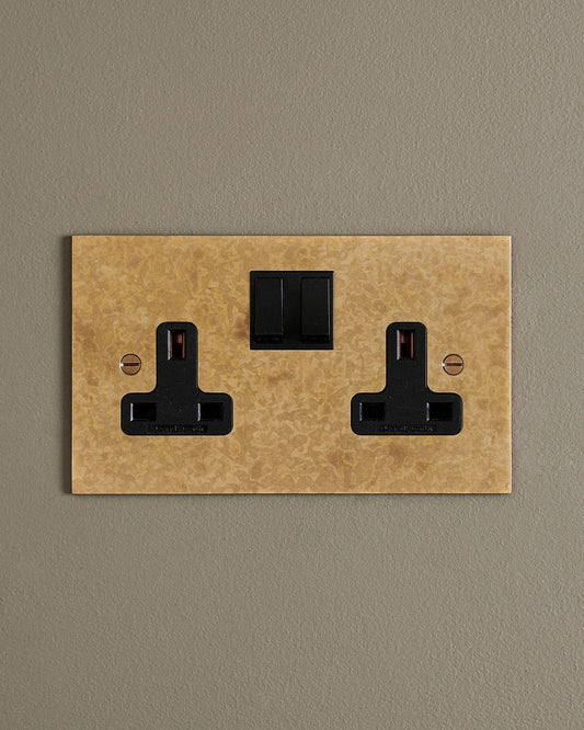 A straight-on view of a classic 13 amp twin plug socket in a mottled aged brass finish with black plastic switches.