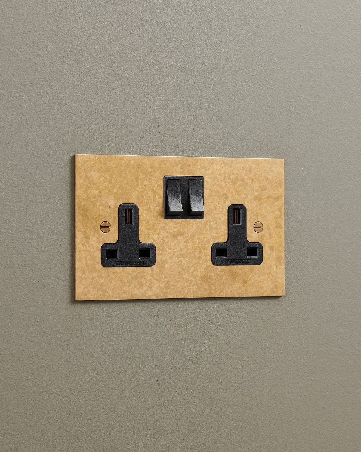 An oblique view of a classic 13 amp twin plug socket in a mottled aged brass finish with black plastic switches. TEST