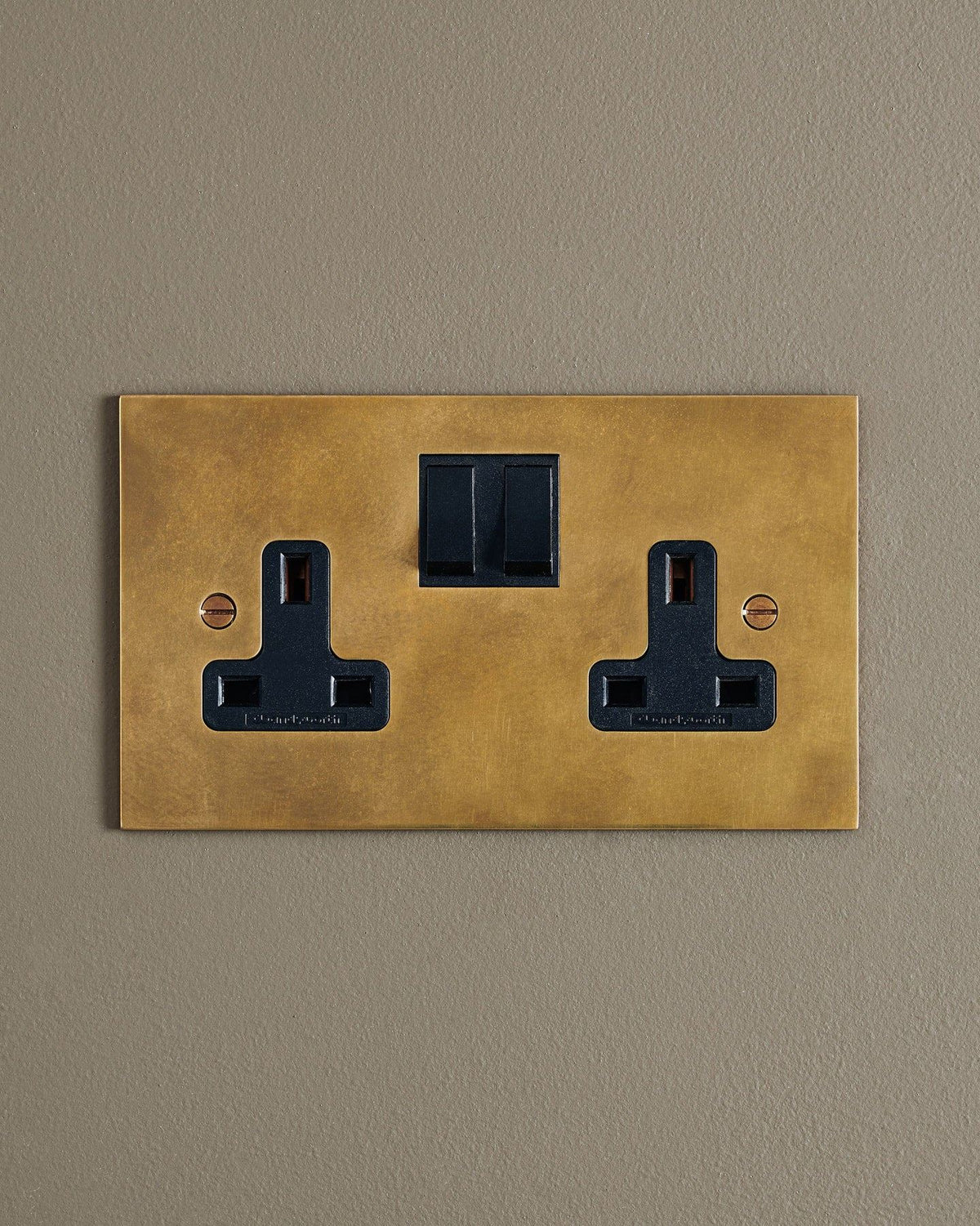 A straight-on view of a classic 13 amp twin plug socket in a warm burnished brass finish with black plastic switches. TEST