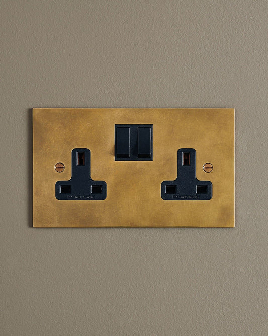 A straight-on view of a classic 13 amp twin plug socket in a warm burnished brass finish with black plastic switches.