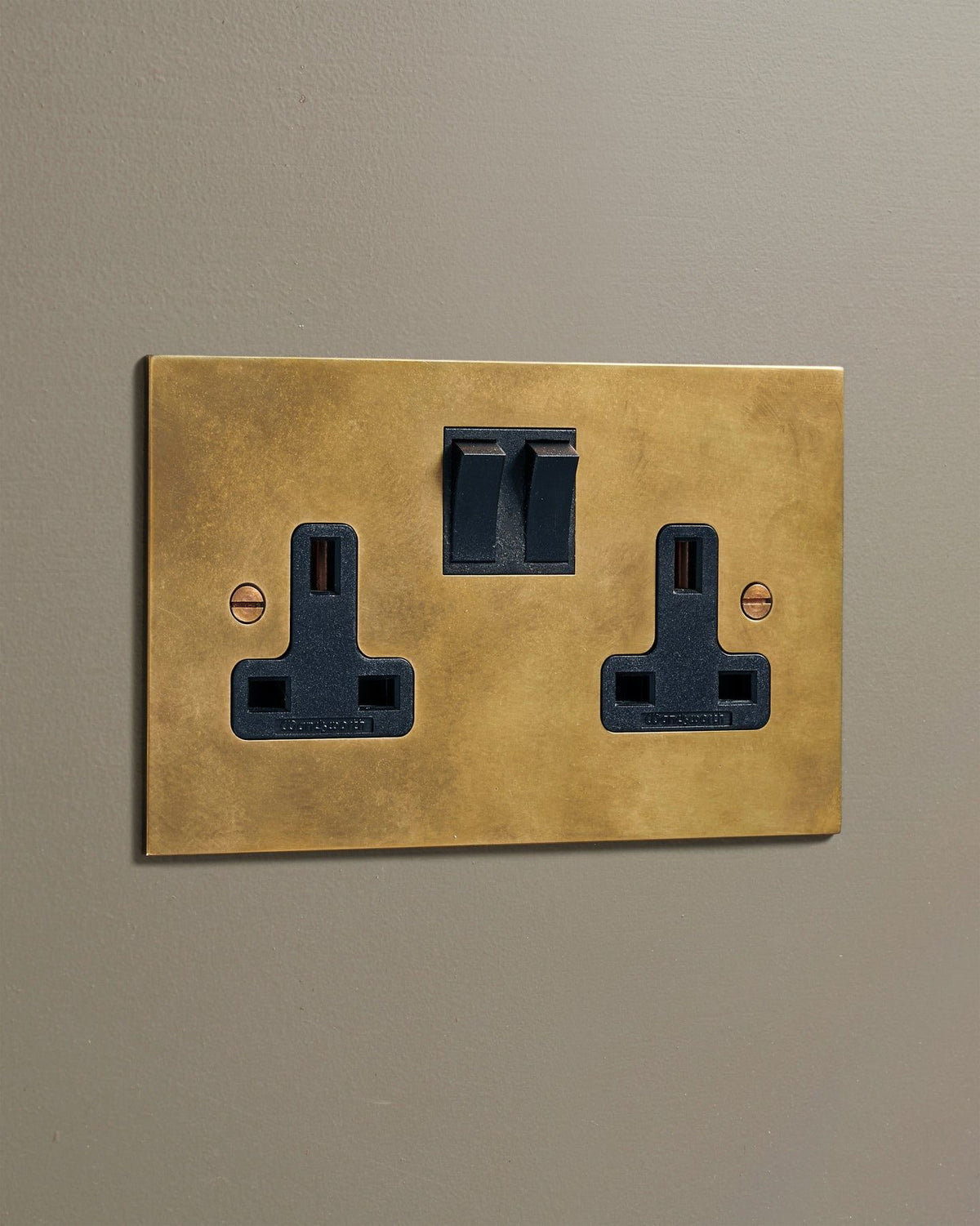 An oblique view of a classic 13 amp twin plug socket in a warm burnished brass finish with black plastic switches. TEST