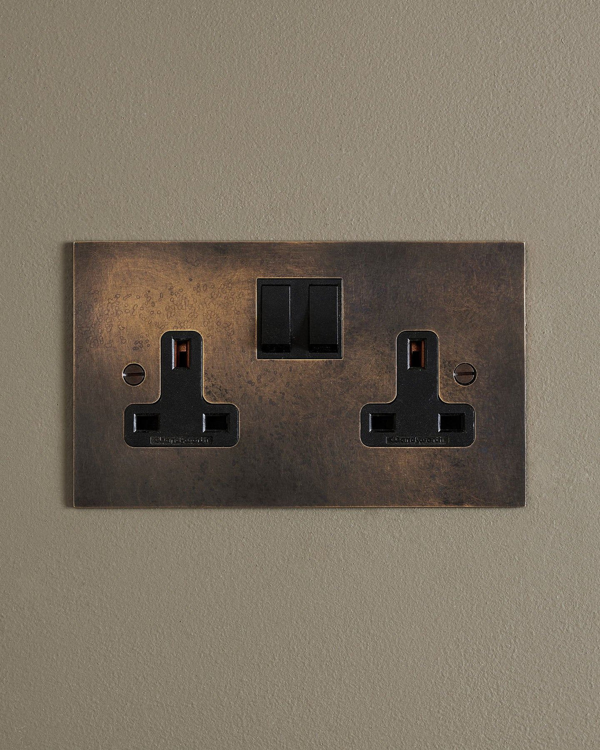 A straight-on view of a classic 13 amp twin plug socket in a dark oxidised brass finish with black plastic switches. TEST