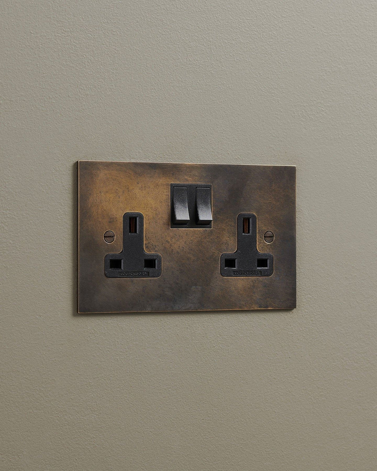 An oblique view of a classic 13 amp twin plug socket in a dark oxidised brass finish with black plastic switches. TEST