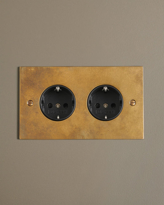 A straight-on view of a classic Schuko-style twin plug socket in a warm burnished brass finish.