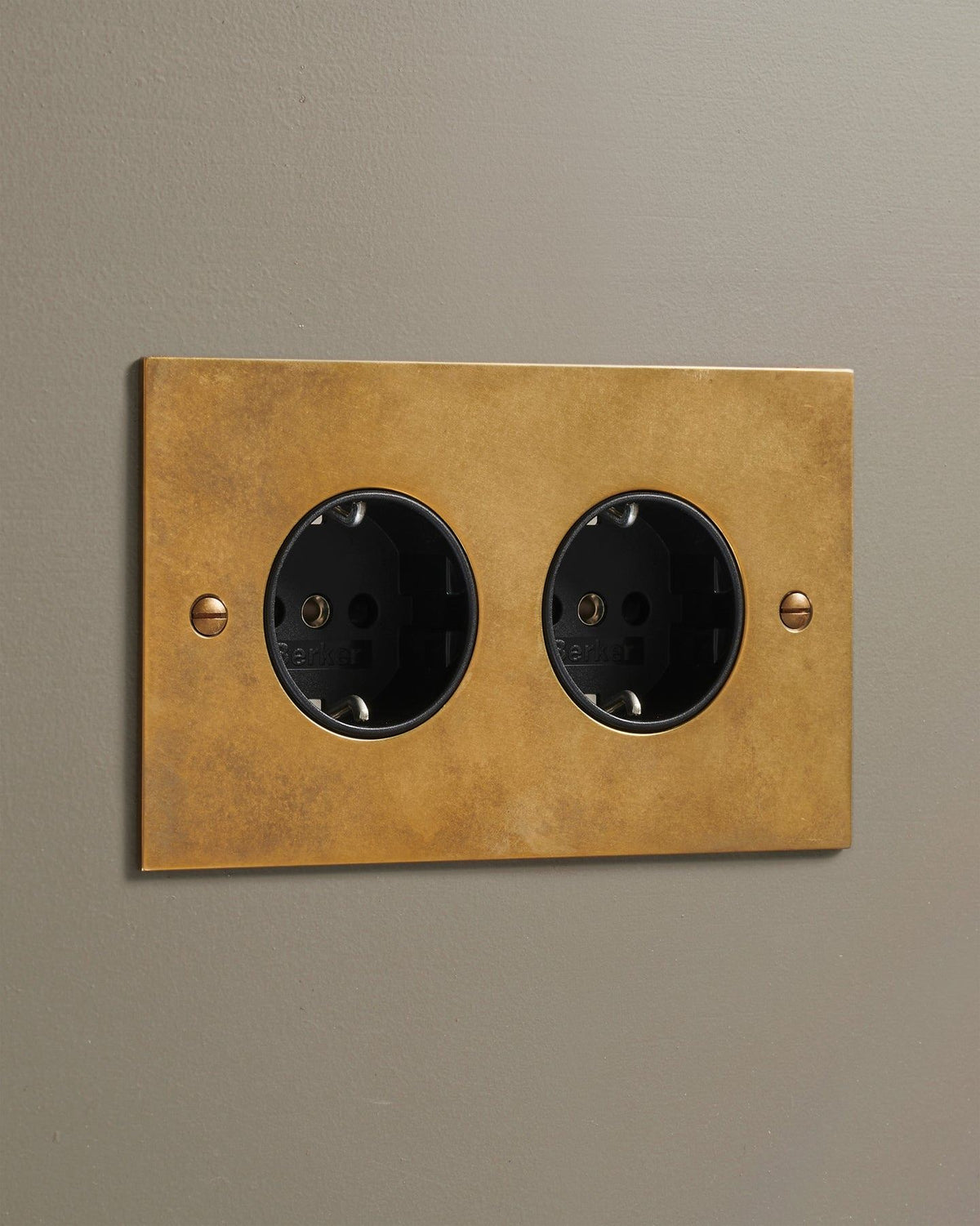 An oblique view of a classic Schuko-style twin plug socket in a warm burnished brass finish. TEST