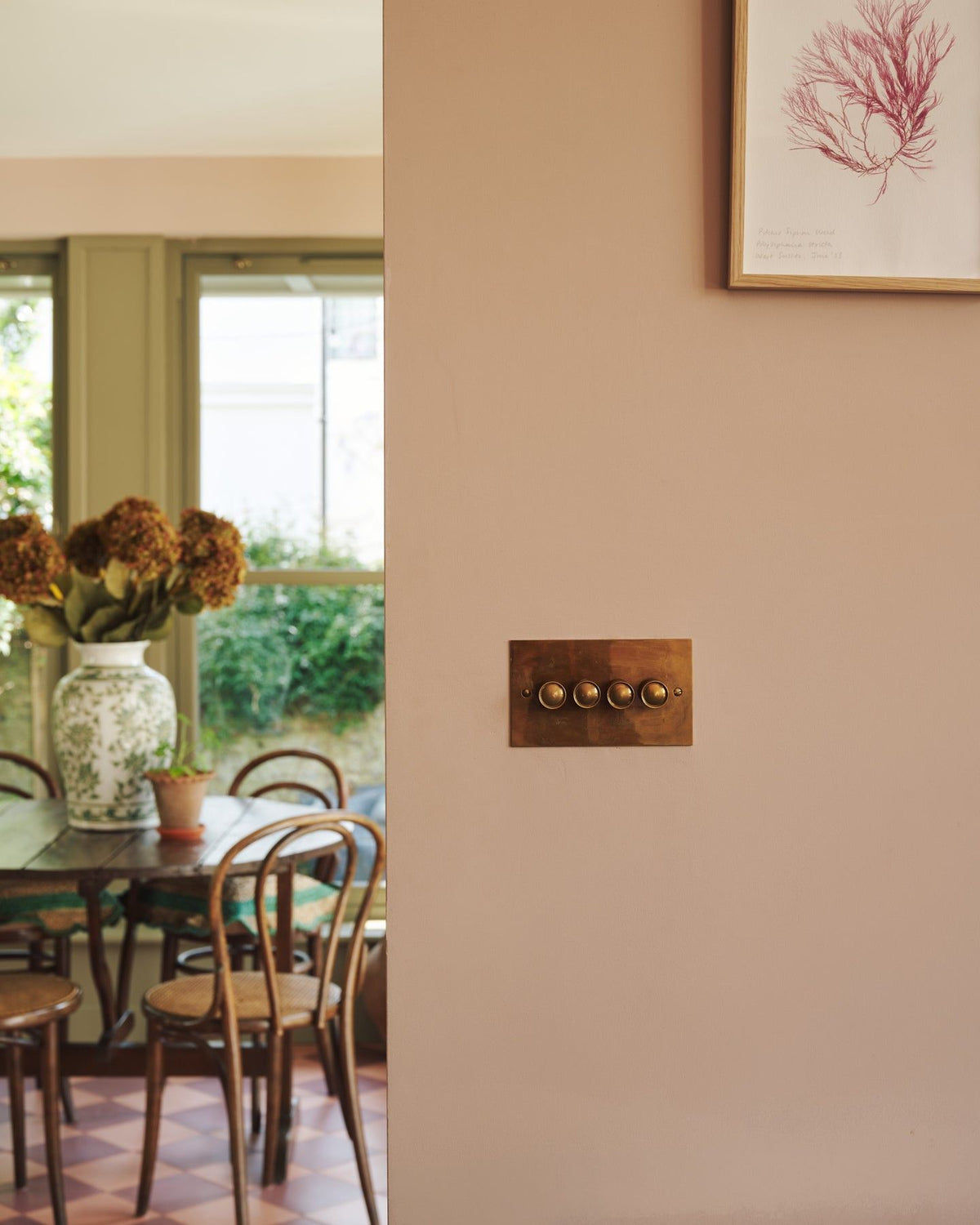 A straight-on view of a 4 gang classic dimmer switch in a warm burnished brass finish on soft pink walls, a cafÈ-style table and chairs with a vase full of pink hydrangeas can be seen in the background. TEST