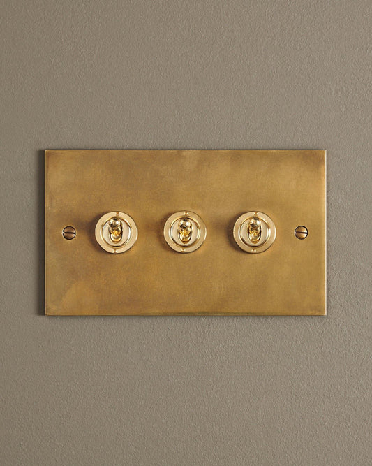 A straight-on view of a 3 gang classic toggle switch in a warm burnished brass finish.