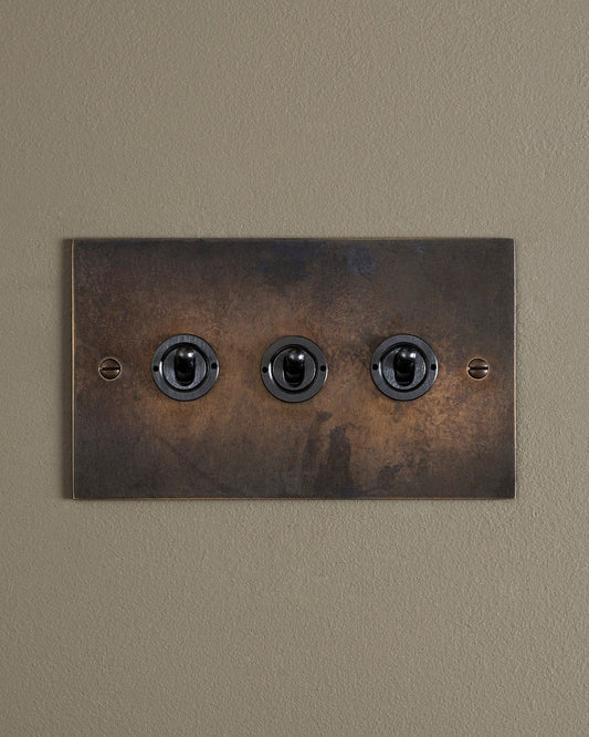 A straight-on view of a 3 gang classic toggle switch in a dark oxidised brass finish.