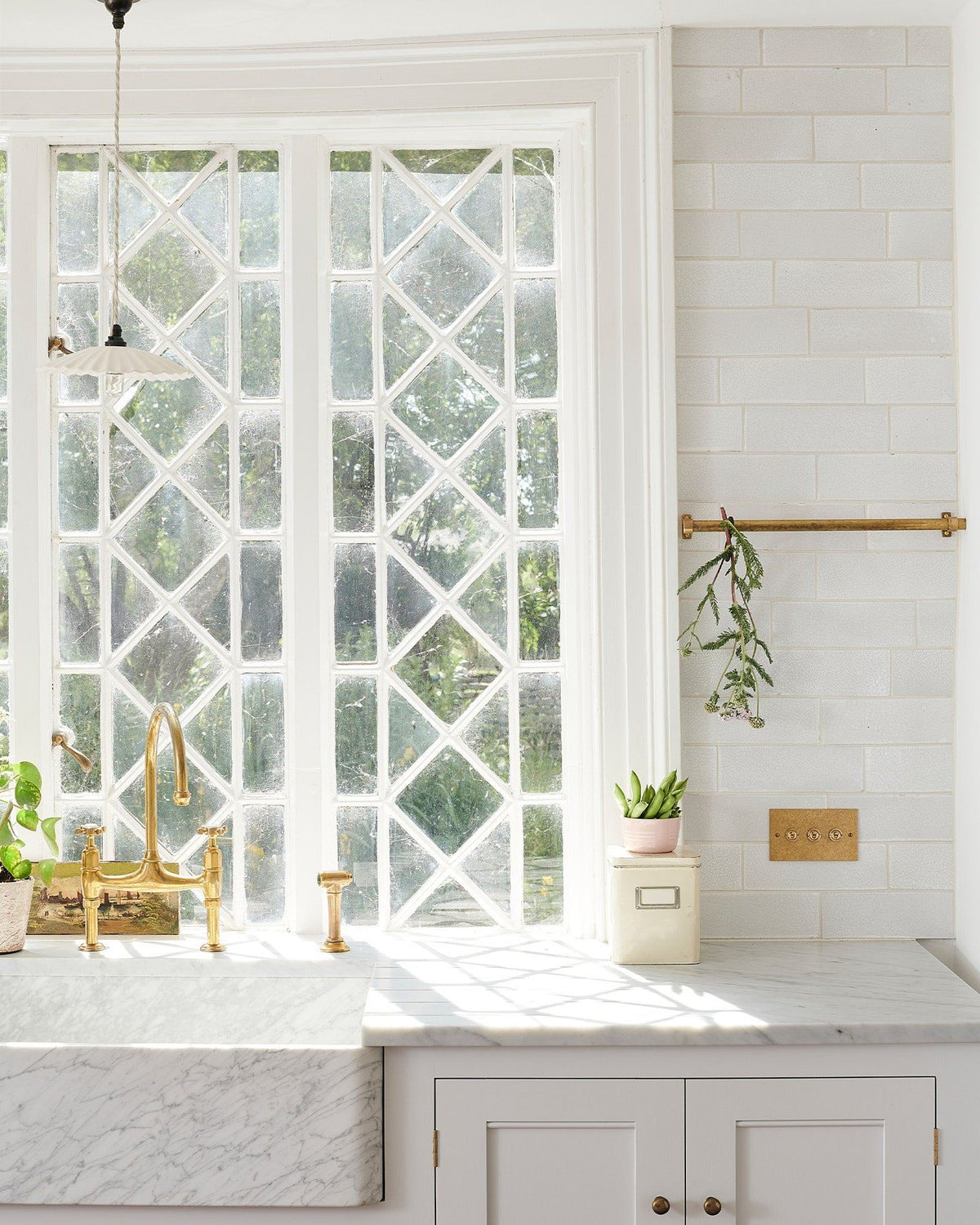 A straight-on view of a 3 gang classic toggle switch in an aged brass finish in a Mediterranean-style kitchen with white painted cupboards, Carrara marble worktop, white metro tiles and original Victorian white leaded windows. TEST