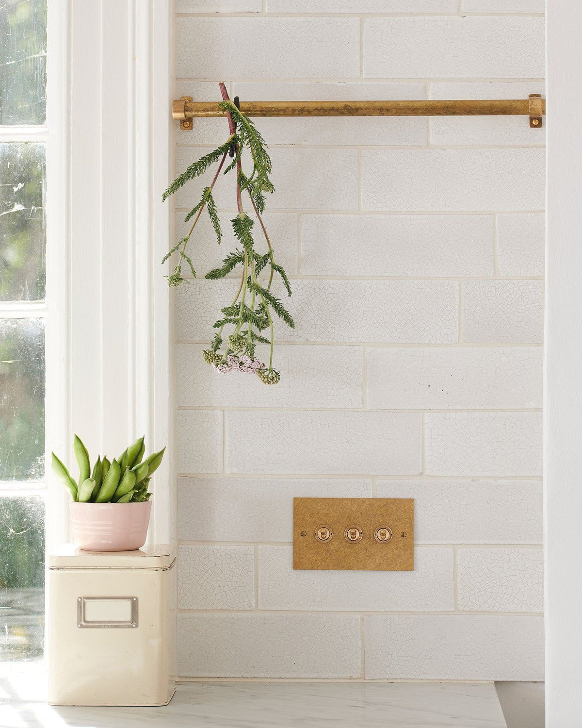 A straight-on view of a 3 gang classic toggle switch in an aged brass finish on white metro tiles with a small olive branch dangling from a short aged brass hanging rail above. TEST
