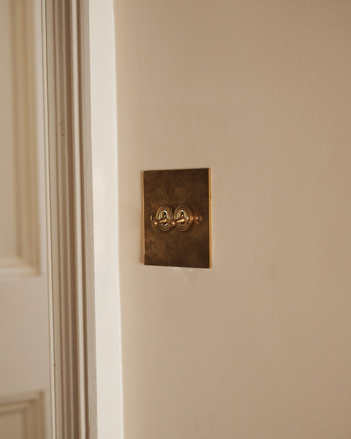 An oblique view of a 2 gang classic toggle switch in a warm burnished brass finish on soft pink walls with light painted woodwork. | 2 Gang shown TEST