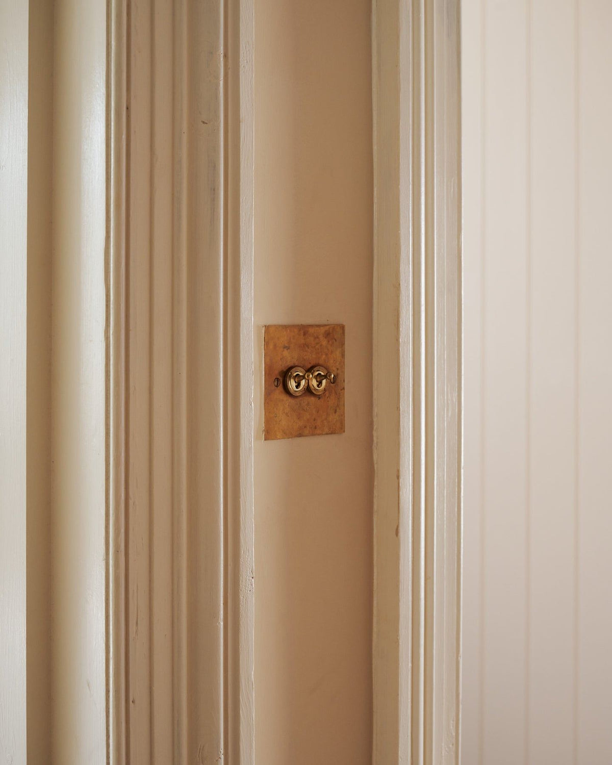 An oblique view of a 2 gang classic toggle switch in a warm burnished brass finish on soft pink walls between traditional light painted architrave. TEST