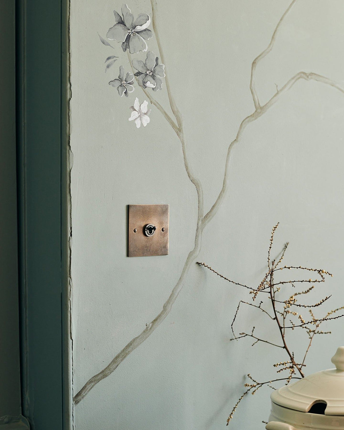 An oblique view of a 1 gang classic toggle switch in a dark oxidised brass finish on a duck-egg blue wall with a delicate handpainted mural of winding branches. TEST