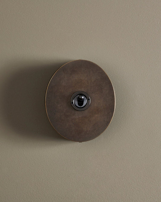 A straight-on view of a 1 gang oval-shaped toggle switch in a dark oxidised brass finish with a black base.