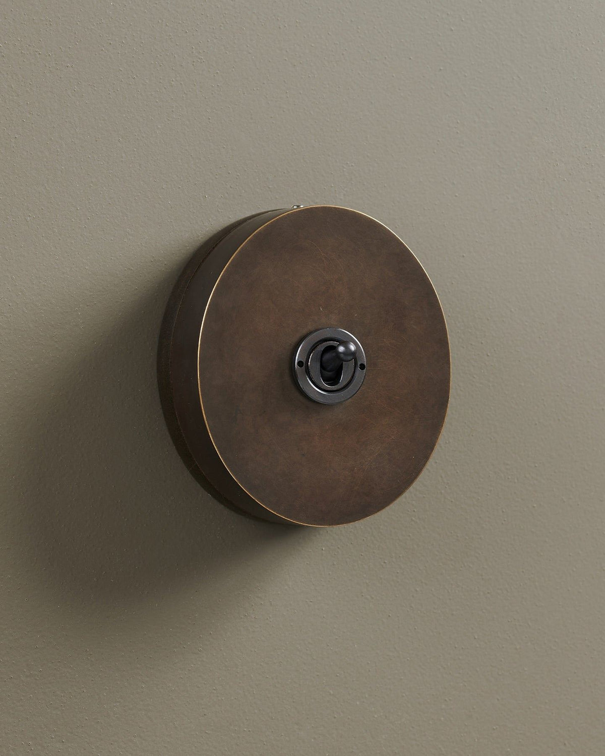 An oblique view of a 1 gang oval-shaped toggle switch in a dark oxidised brass finish with a black base. TEST