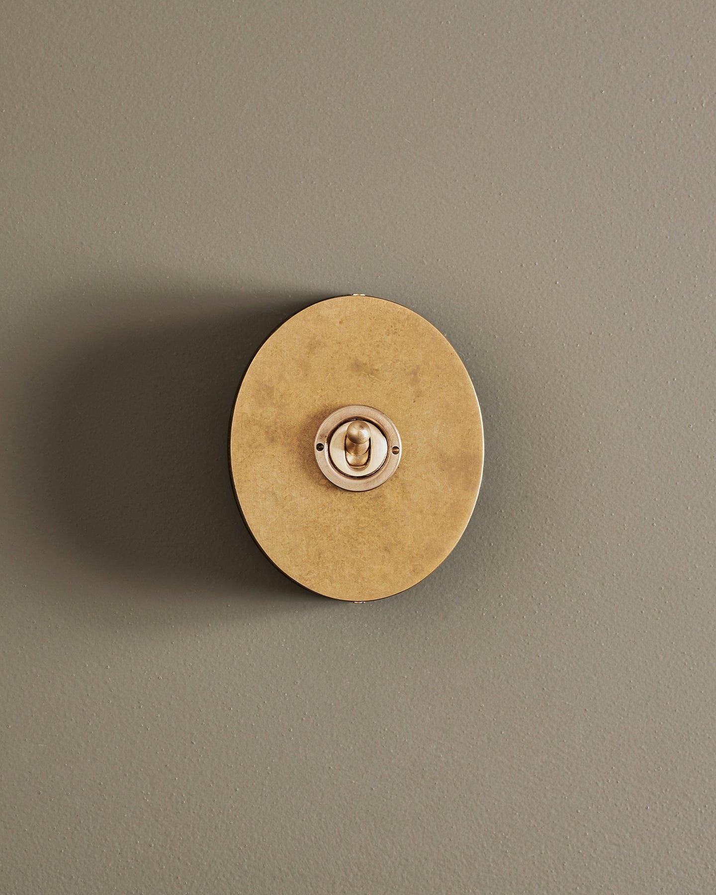 A straight-on view of a small 1 gang oval-shaped toggle switch in a mottled aged brass finish with a black base.