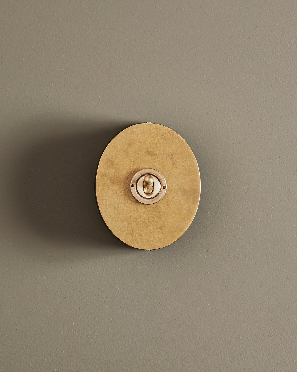 A straight-on view of a small 1 gang oval-shaped toggle switch in a mottled aged brass finish with a black base. TEST
