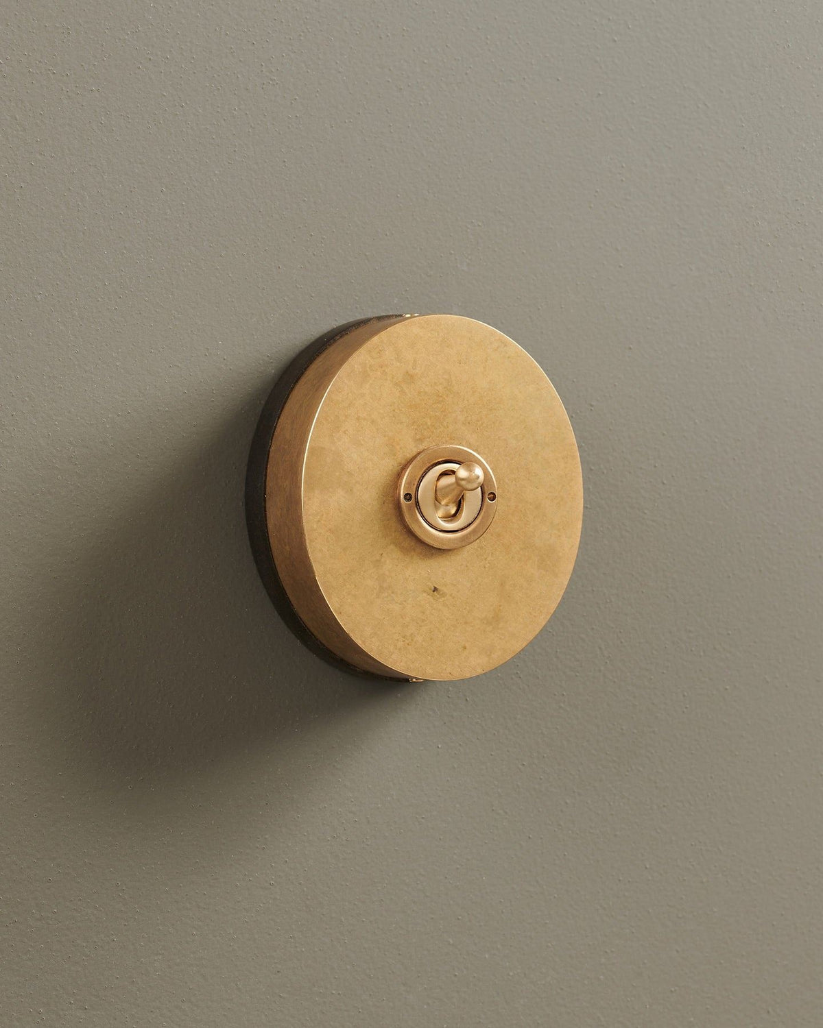 An oblique view of a small 1 gang oval-shaped toggle switch in a mottled aged brass finish with a black base. TEST