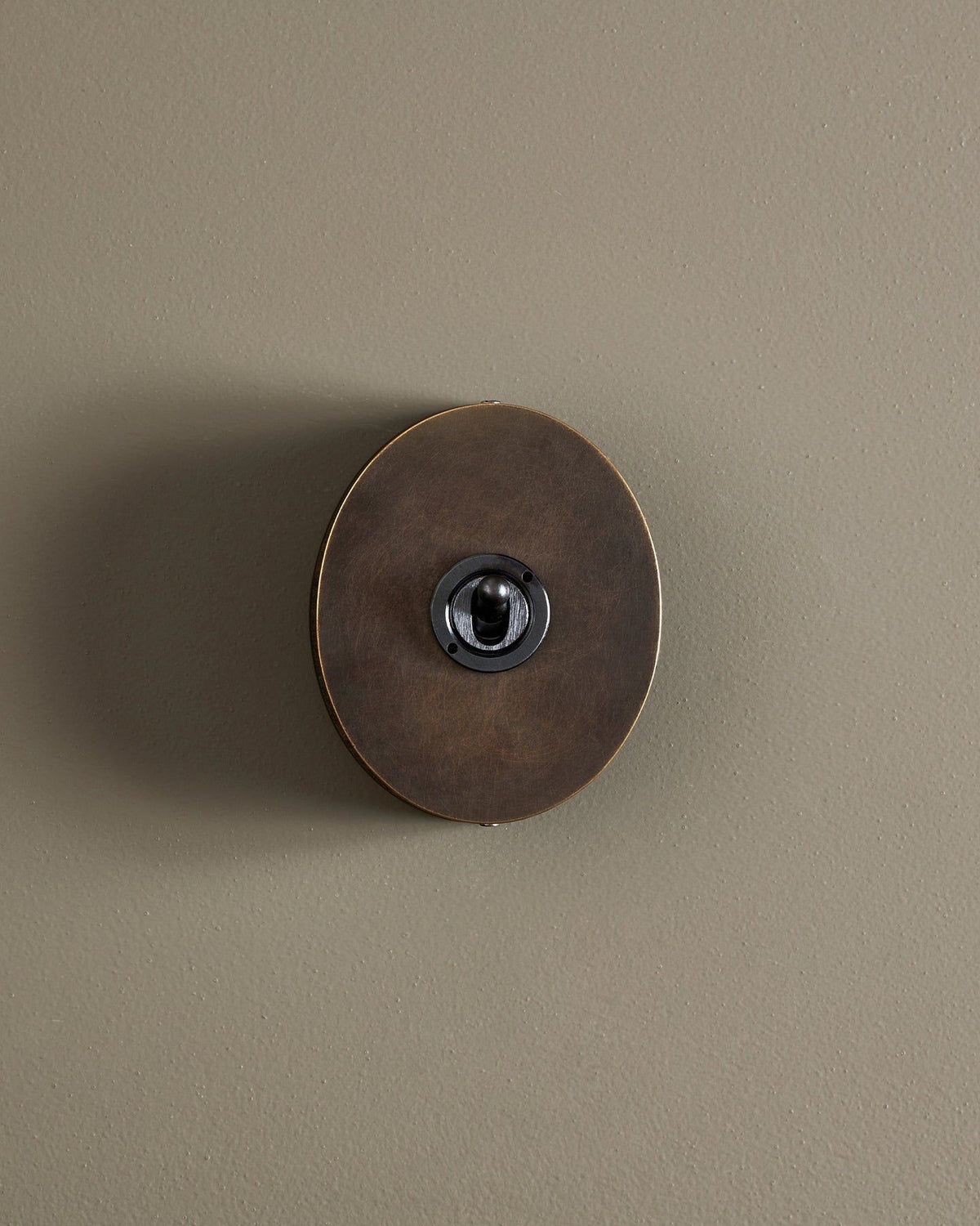 A straight-on view of a small 1 gang oval-shaped toggle switch in a dark oxidised brass finish with a black base. TEST