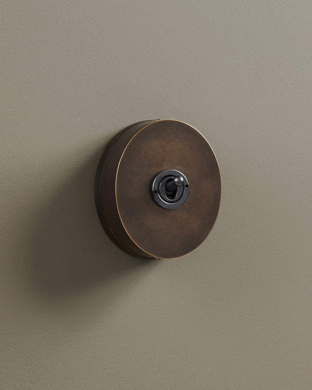 An oblique view of a small 1 gang oval-shaped toggle switch in a dark oxidised brass finish with a black base. TEST