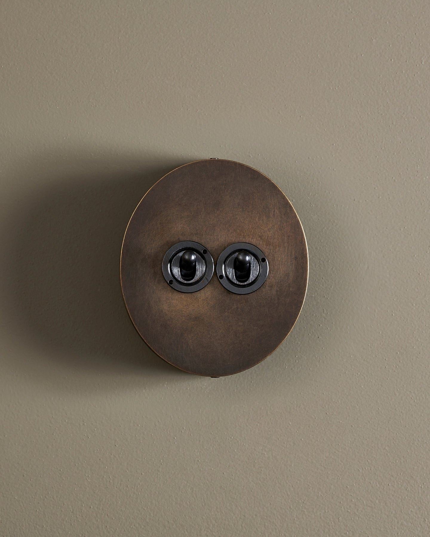 A straight-on view of a 2 gang oval-shaped toggle switch in a dark oxidised brass finish with a black base.