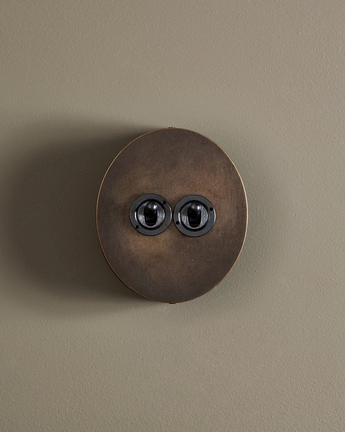 A straight-on view of a 2 gang oval-shaped toggle switch in a dark oxidised brass finish with a black base. TEST