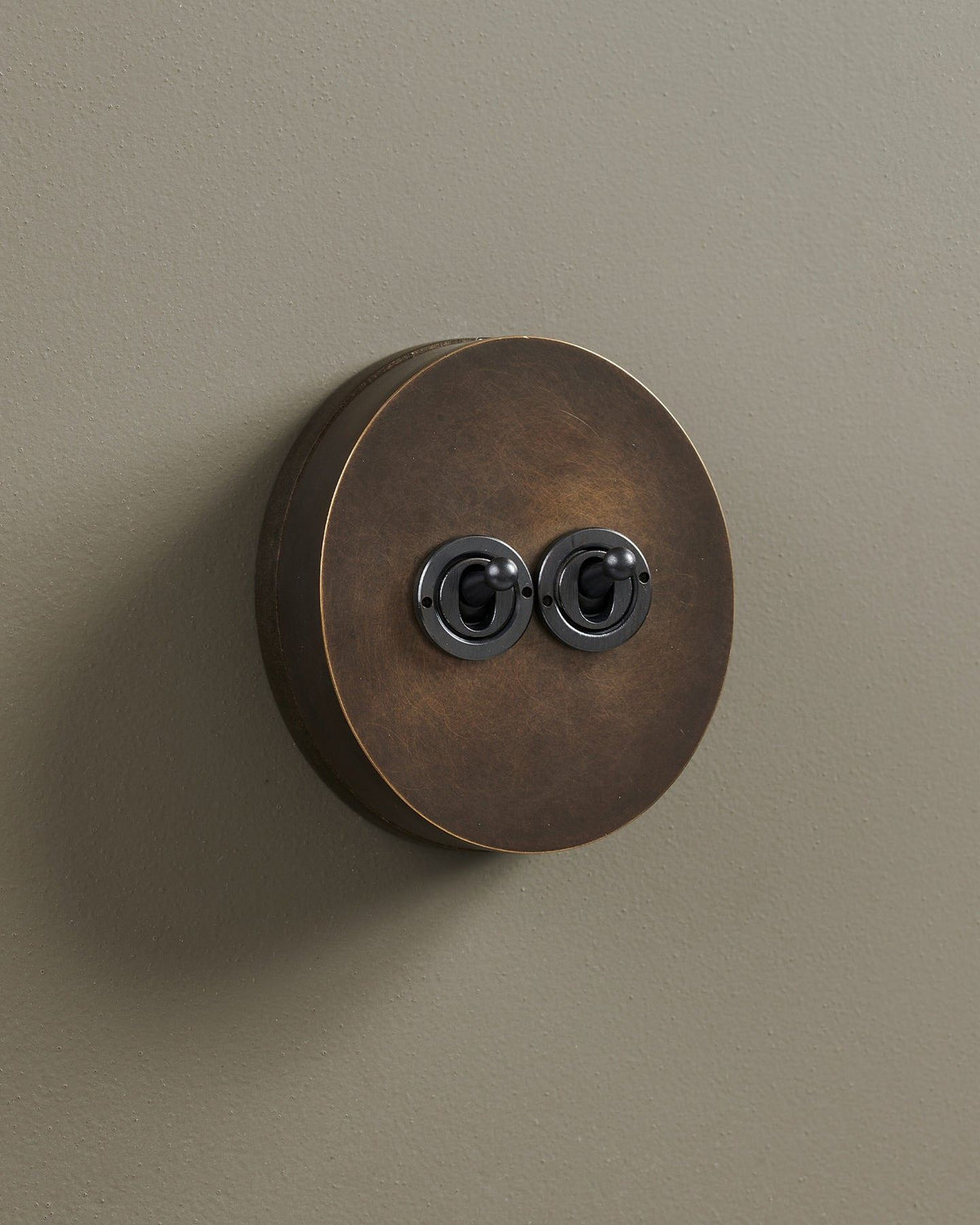 An oblique view of a 2 gang oval-shaped toggle switch in a dark oxidised brass finish with a black base. TEST