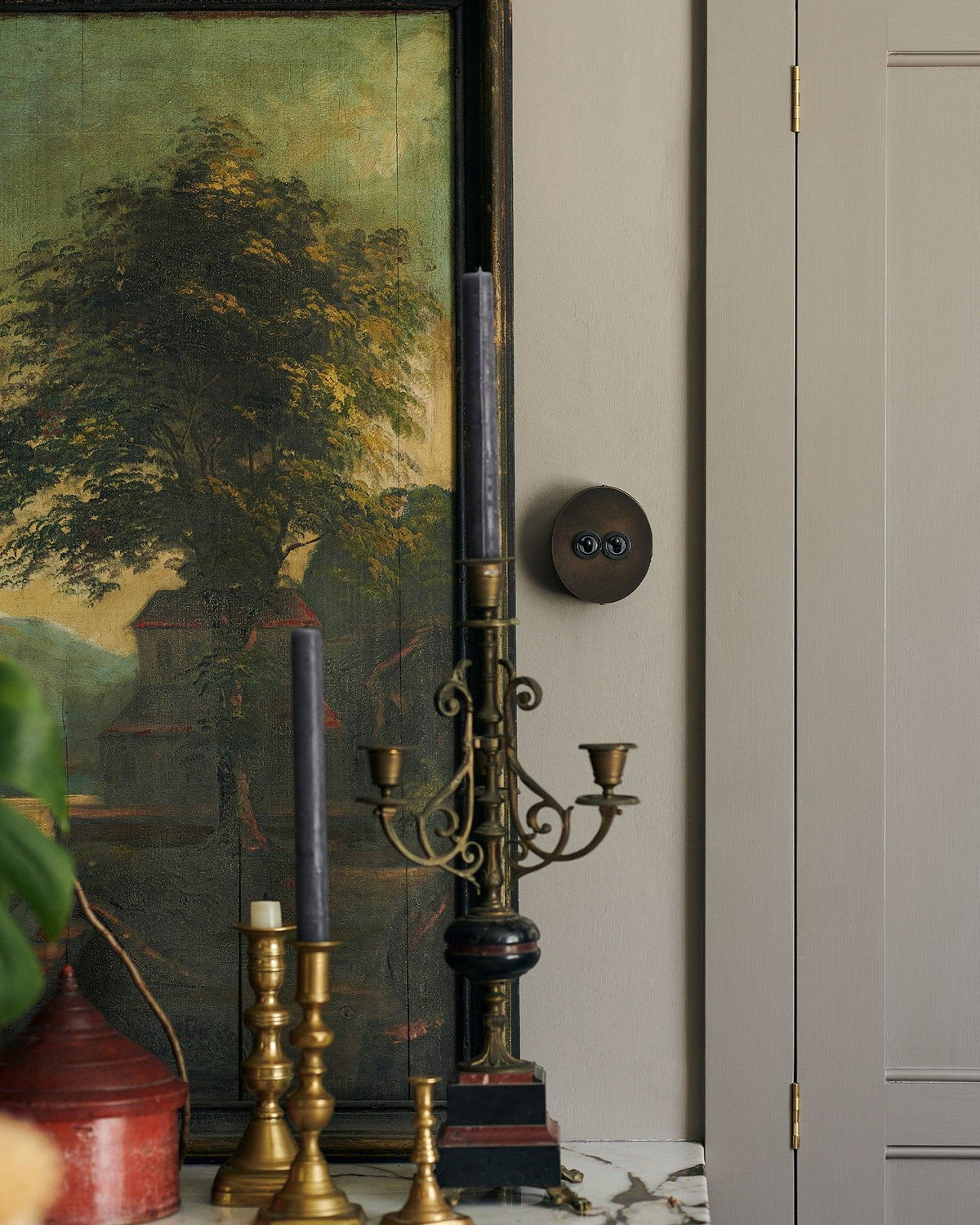 A 2 gang oval-shaped toggle switch in a dark oxidised brass finish on a grey wall, beside a large antique oil painting of a moody landscape. An elaborate tall candelabra sits in the foreground. TEST