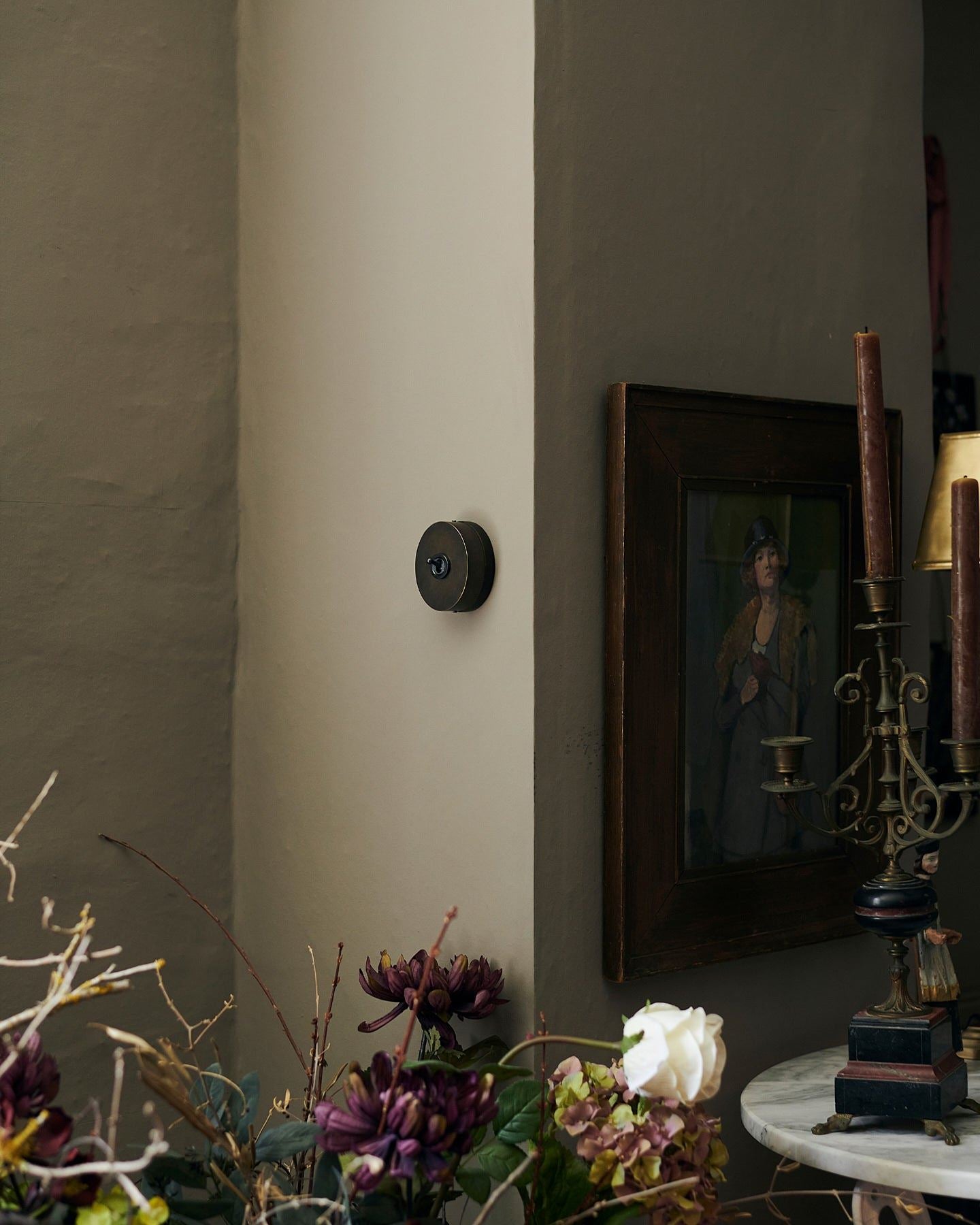 A small 1 gang oval-shaped toggle switch in a dark oxidised brass finish on a grey wall, a moody impressionist oil painted hangs on the wall around a corner with dried flowers in the foreground.