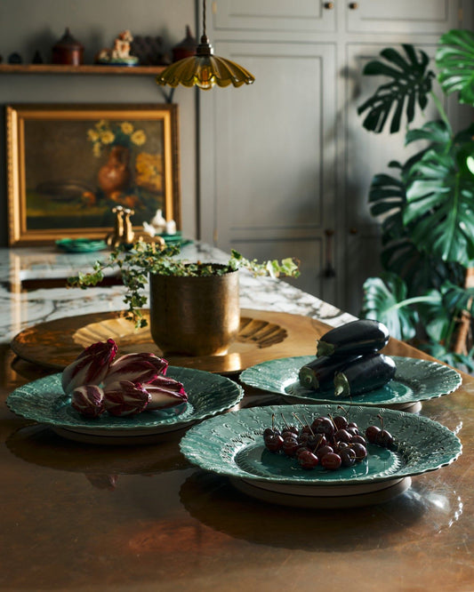 Three large handmade bohemian-style green ceramic platters with an assortement of aubergines, red chicory and cherries in an eclectic kitchen with an aged copper worktop, grey painted walls, antique paintings and a large monstera plant.