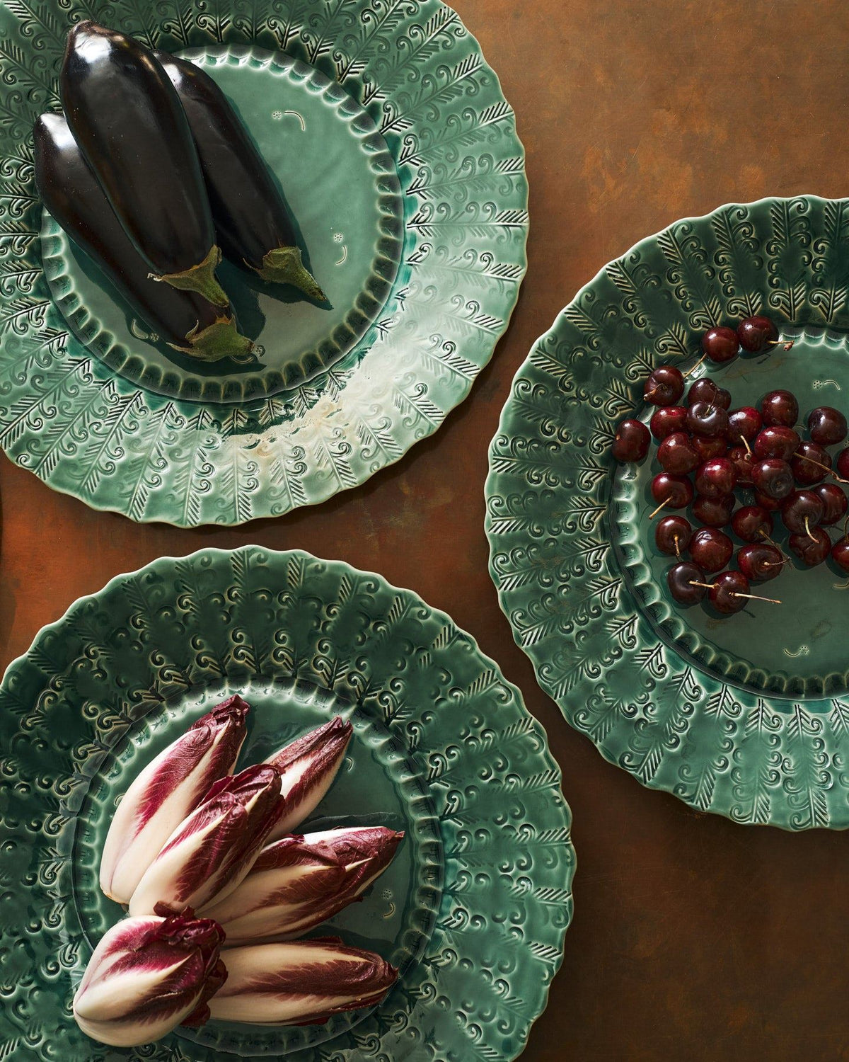 An overhead view of three large handmade bohemian-style green ceramic platters with scalloped rims and delicate impressions and surface patterns and an assortement of aubergines, red chicory and cherries on an aged copper worktop. TEST