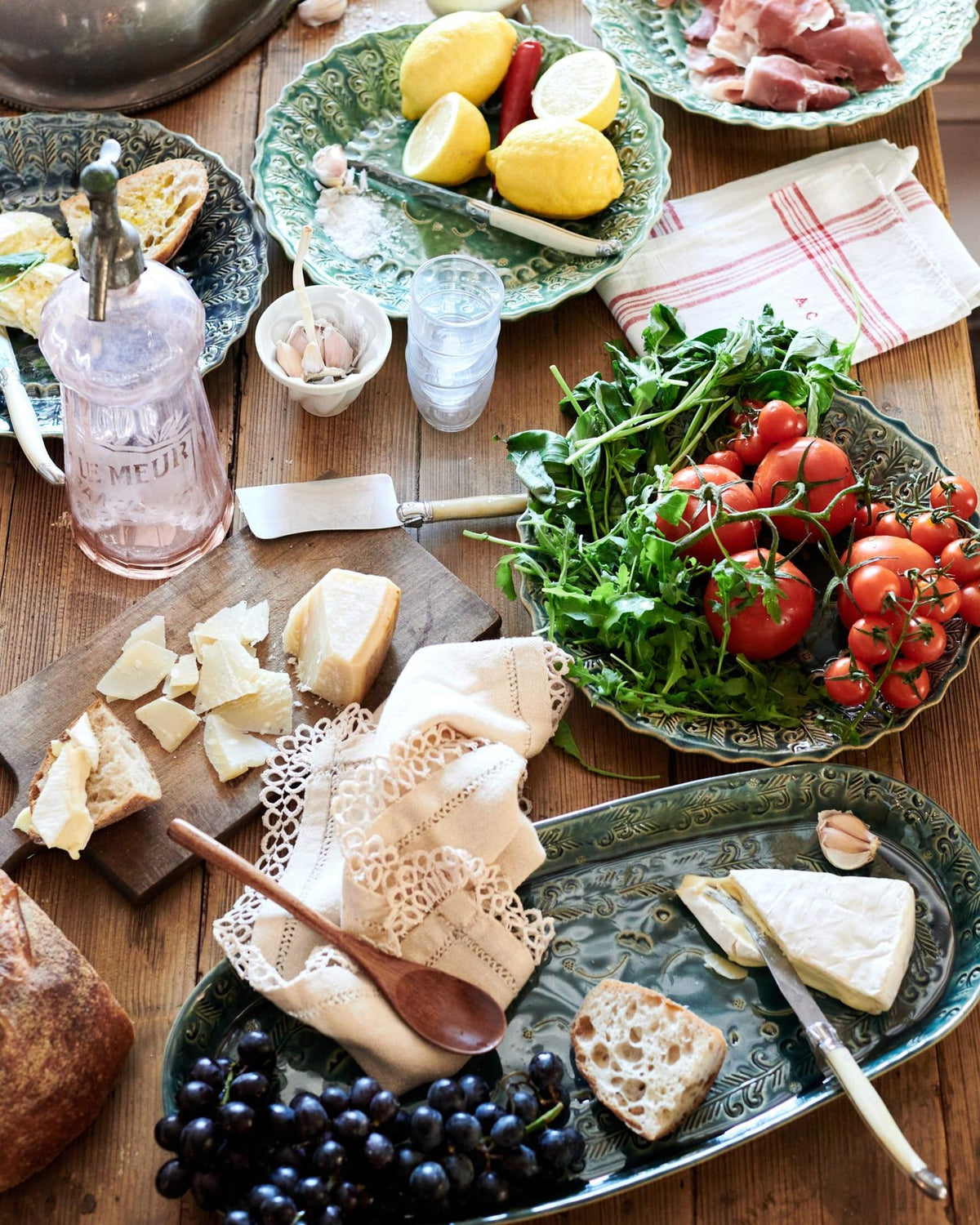Several oval and round handmade bohemian-style ceramic platters, with black grapes, cheeses, homemade bread, lemons, chillis, prosciutto ham, tomatoes and leafy greens. TEST